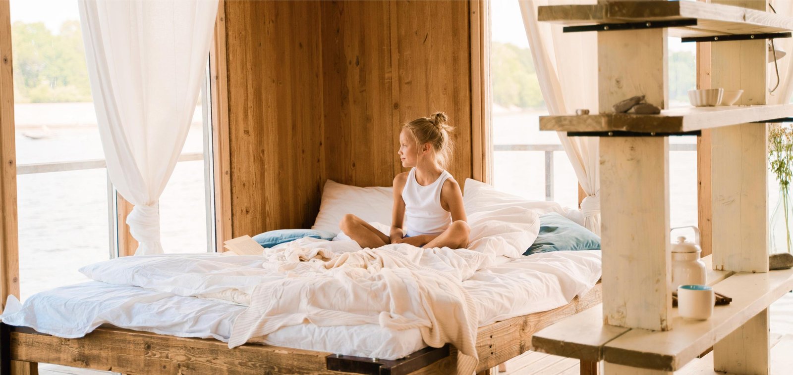 Peaceful Morning in a Rustic Bedroom Young girl sitting on a bed in a wooden room with white curtains and a view of water.
