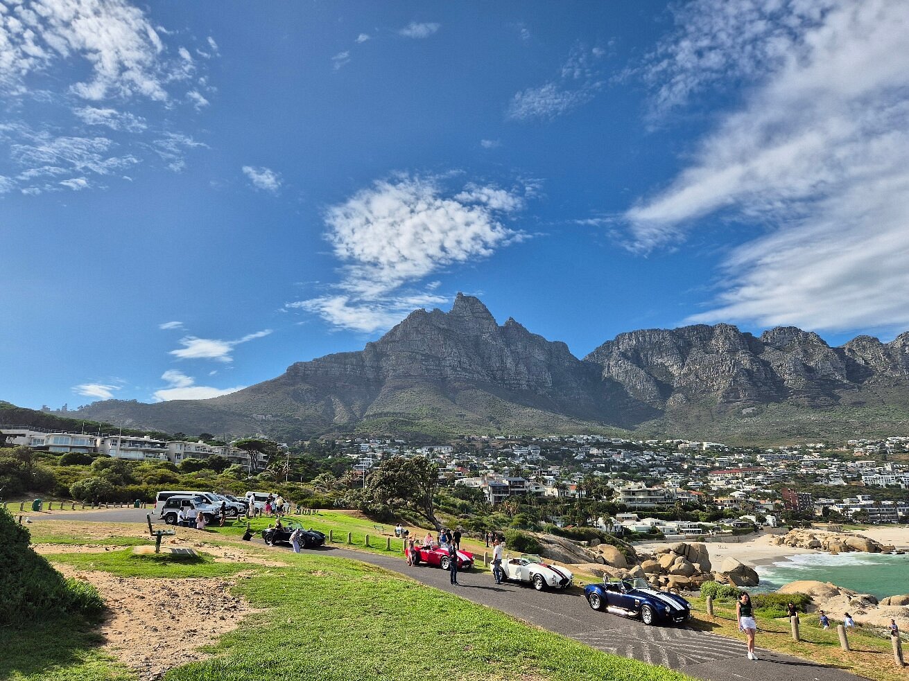 Classic cars and Cobra replicas parked on a scenic road in Cape Town with the Twelve Apostles mountains and beach.